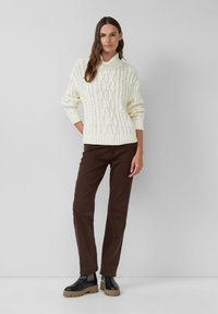 Femme portant un pull à col haut en tricot crème, un pantalon droit marron foncé et des bottes noires à semelle épaisse, se tenant devant un mur uni.
