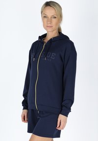 Navy zip-up hoodie made of soft fabric, featuring a gold zipper, front pocket, and a matching shorts set. Logo text displayed on the chest.