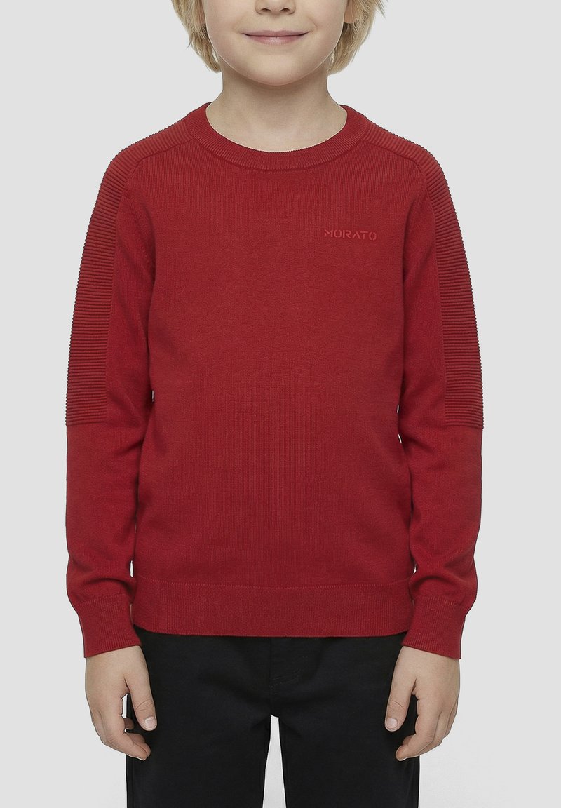 Enfant portant un pull rouge à manches longues avec des détails côtelés aux épaules et un pantalon noir, debout devant un fond clair uni.