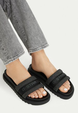 Piedi che indossano sandali a ciabatta G-Star RAW neri e grigi con smalto bianco sulle unghie dei piedi e jeans grigi slavati su uno sfondo bianco.