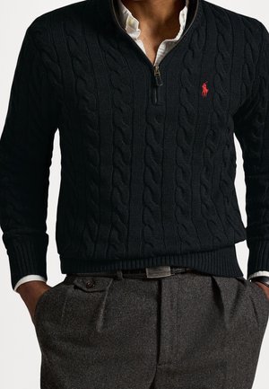 Hombre con jersey oscuro de punto trenzado y cremallera corta con logotipo rojo sobre camisa blanca, combinado con pantalones grises plisados y cinturón negro.