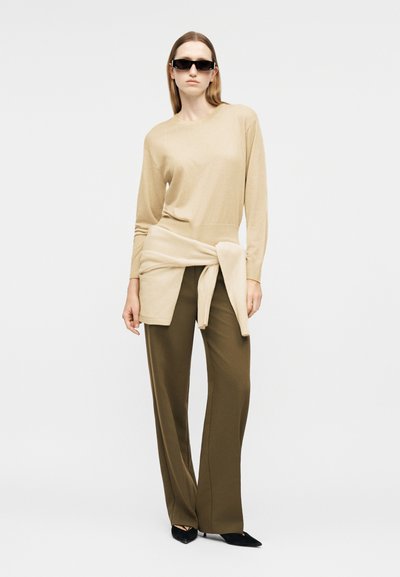 Femme portant un haut à manches longues beige, un pull assorti noué à la taille, un pantalon large olive, des chaussures pointues noires et des lunettes de soleil noires, debout.