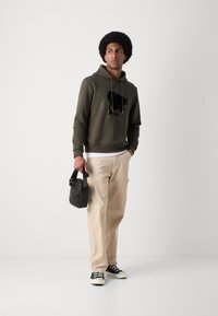 Sweat à capuche vert olive avec des détails graphiques, pantalon beige et baskets noires. Tient un petit sac noir avec une sangle.