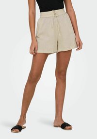 Shorts en lin beige avec une taille élastique à cordon, des poches latérales et une coupe décontractée. Assortis à des sandales glissantes noires.