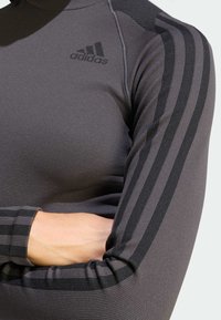 Čierne športové tričko s dlhým rukávom s rebrovanou textúrou a tromi čiernymi pruhmi na rukávoch, s vyrazeným logom na hrudi.