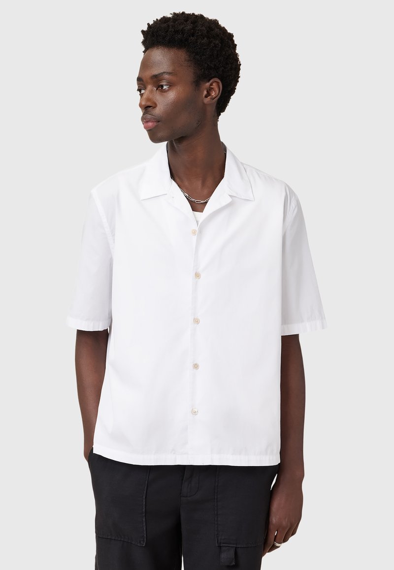 Camisa blanca de manga corta, con botones en el frente, cuello extendido, corte relajado, fabricada en un suave algodón, presentada sobre un fondo neutro.