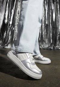 Silver sneakers with white accents, smooth leather upper, perforated toe box, and a rounded sole, featured against a textured metallic backdrop.