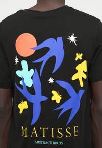 Fekete pamut póló színes absztrakt madarakkal, növényzettel és égi elemekkel. Az alján sárga "MATISSE" felirat, alatta pedig az "ABSTRAKT MADARAK" aláhúzva.