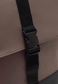 Détail d'un sac imperméable gris avec une sangle noire réglable et une boucle en plastique, présentant des coutures visibles sur le bord. Texture lisse.