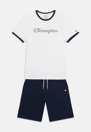 Camiseta de algodón blanca con ribete azul marino y logo de "Champion", combinada con pantalones cortos azul marino con bolsillos laterales y cordón ajuste.