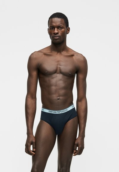 Άνδρας μοντέλο με σκούρο δέρμα, κοντά σγουρά μαλλιά, φορώντας σκουρό μπλε εσώρουχα Emporio Armani, στέκεται μπροστά σε ένα απλό φωτεινό φόντο.