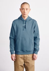 Sweatshirt azul de algodão com um bolso frontal, capuz ajustável e logótipo branco. O modelo veste calças caqui e está posicionado contra um fundo neutro.