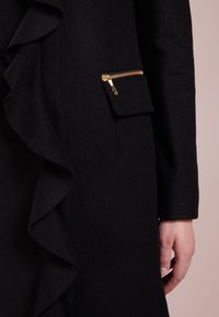 Cappotto nero con bordo arricciato e tasca con zip in metallo dorato. Realizzato in un tessuto testurizzato, presenta una silhouette aderente.