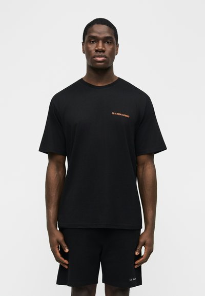 Νέος άνδρας που στέκεται προς τα εμπρός φορώντας μαύρο T-shirt με μικρό κείμενο "LES BENJAMINS" και ταιριαστή μαύρη βερμούδα.