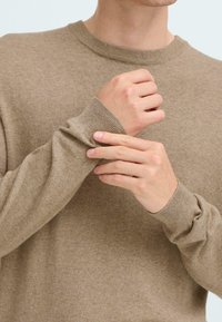 Maglione beige a maglia con scollatura rotonda. La foto ravvicinata mostra il tessuto texturizzato e i polsini aderenti, mani che sistemano la manica.
