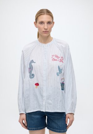 Kvinde iført en hvid stribet bluse med broderi af en søhest, svamp, fugl, flaske og rød tekst med teksten "I Run My Own Show."