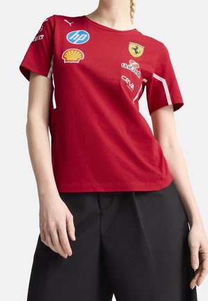 Osoba ubrana w czerwoną koszulkę Ferrari z logo sponsorów, w tym HP, Shell, UniCredit i CEVA, w połączeniu z czarnymi spodniami.
