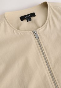Beige rits-top met een ronde halslijn, voorzien van een metallic ritsdetail aan de voorkant en een gladde, zacht aanvoelende stof.