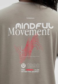 Rückseite eines beigen Shirts mit den Aufschriften "lululemon", "Mindful Movement", einem roten wellenförmigen Diagramm, "1998" und "Finde den Fluss auf der Reise".