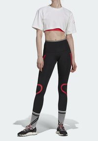 Biały skrócony t-shirt, czerwony stanik sportowy pod spodem, czarne legginsy z czerwonymi akcentami oraz czarne buty sportowe w pasiastych skarpetkach.