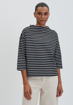 Vrouw met donker haar in een paardenstaart draagt een zwart-wit horizontaal gestreept shirt met driekwart mouwen en lichtbeige broek.