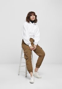Pullover bianco con cappuccio e design arricciato, abbinato a pantaloni cargo marroni con tasche e sneakers bianche con suola texturizzata.