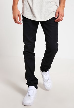 Muž v úzkých černých džínách a bílých teniskách, chodící s viditelnýma rukama a dolní částí trupu.