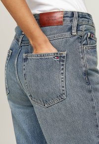 Pantalones vaqueros de mezclilla en azul claro con un ligero desgaste y una etiqueta de cuero de Tommy Hilfiger en la cintura. Presentan bolsillos traseros estándar.