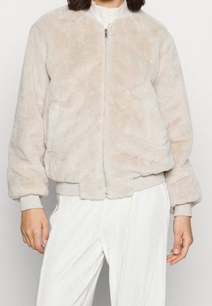Kvinde iført en lysebeige fluffy lynlåstrøje med ribstrikkede manchetter over hvide ribstrikkede bukser mod en ensfarvet baggrund.