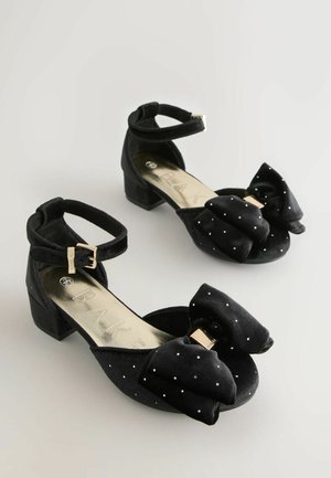 Schwarze Stoffschuhe mit einem niedrigen Blockabsatz, ausgestattet mit einer großen Schleife, silbernen Punkten und einem Knöchelriemen mit einer goldenen Schnalle.