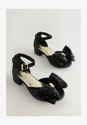 Czarne buty z materiału z niskim obcasem w kształcie bloku, z dużą kokardą, srebrnymi kropkami oraz kostką z zapięciem na złotą klamrę.