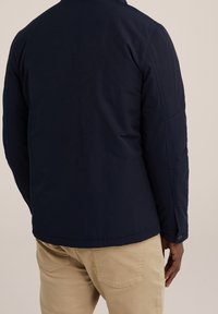 Man i marinblå jacka och beige byxor, sedd bakifrån mot en enkel ljus bakgrund.