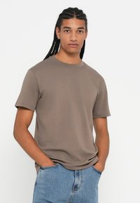 Young man with long braided hair wears a taupe short-sleeve t-shirt and light blue jeans, standing with hands in pockets against white background.