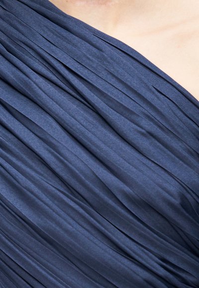 Gros plan sur le haut du torse et l'épaule d'une personne portant un vêtement en tissu plissé bleu foncé.