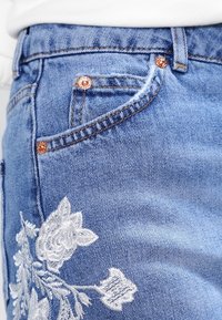 Světlé modré džíny z denimu s detailním bílým květinovým výšivem na levém stehně, vybavené měděným kováním a viditelným prošitím.