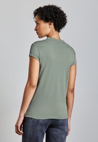 Zelené, krátké rukávy, přiléhavé tričko s kulatým výstřihem. Vyrobeno z měkké tkaniny s hladkou texturou. Nenošené s tmavě šedými džínami.