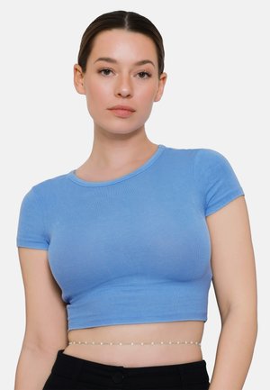 Młoda kobieta o ciemnych włosach związanych do tyłu nosi dopasowany niebieski krótki top i cienki łańcuszek w pasie na białym tle.