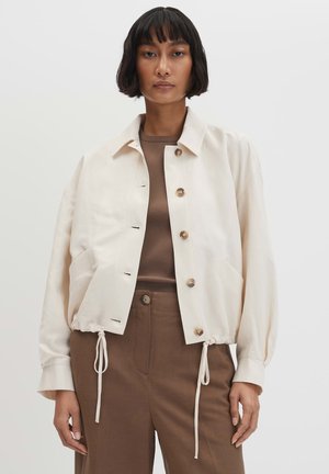 Femme portant une veste crème à boutons avec cordons ainsi qu'une chemise et un pantalon marron côtelés sur fond blanc uni.