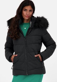 Giubbotto nero imbottito con chiusura a zip, dotato di cappuccio in pelliccia sintetica, design trapuntato e tasche laterali. Indossato sopra una maglia verde.