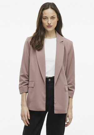 Světle růžový blazer s přiléhavým střihem, se špičatými klopami, dvěma předními kapsami a tříčtvrtečními rukávy, zkombinovaný s bílým topem a černými kalhotami.