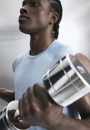 Mladić pletenih kosa podiže srebrnu bučicu, nosi svijetloplavu sportsku majicu, fokusiran na trening snage.