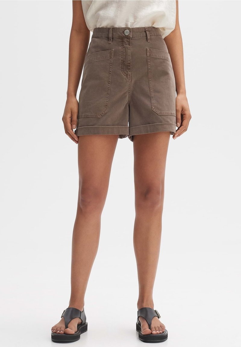 Femme portant un short marron taille haute avec poches avant, un haut blanc et des sandales plates noires, debout devant un fond uni.