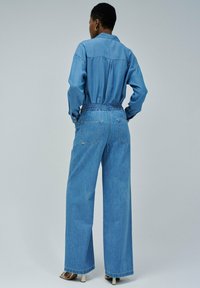 Salopette en denim avec un design de chemise col, taille élastique, pantalons à jambe large et une teinte bleu clair. Poches arrière visibles et détails de couture.