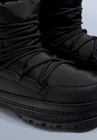 Czarne wodoodporne buty z wyściełaną cholewką, zapinane na krzyżowe sznurowadła oraz z teksturowaną gumową podeszwą zapewniającą przyczepność. Gładki syntetyczny materiał.