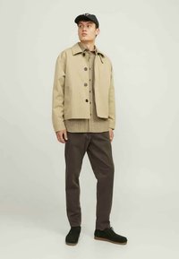 Beige jakke med krave og knaplukning, lagdelt over en mønstret skjorte, kombineret med brune bukser og sorte sneakers. Sort kasket som accent.