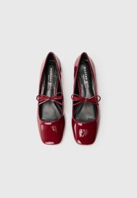 Sapatilhas de balé de verniz burgundy com bicos quadrados, palmilhas pretas e condizes decorativas na parte da frente.