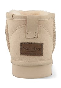 Beige suède laars met een zachte binnenvoering, een gestapelde hak en een geborduurd label "Warmbat Australia" aan de achterkant.