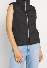 Schwarze ärmellose Puffer-Weste mit hohem Kragen, Reißverschluss vorne, seitlichen Taschen und gestepptem Design. Getragen über hellblauen Jeans.