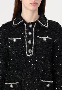 Casaco preto texturizado com lantejoulas, gola adornada, bolsos e detalhes em botões prateados. Detalhado com botões em forma de flor.