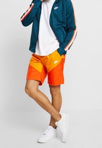 Sportovní bunda Nike v tyrkysové barvě s oranžovými pruhy, doplněná oranžovožlutými plavkami s přechodem odstínů a bílými teniskami.